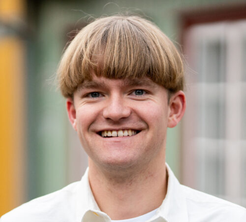 Smilende person med kort, lys blond hår i en hvit, åpenkragen skjorte; bakgrunn med fargerike, utydelige bygg og vinduer.