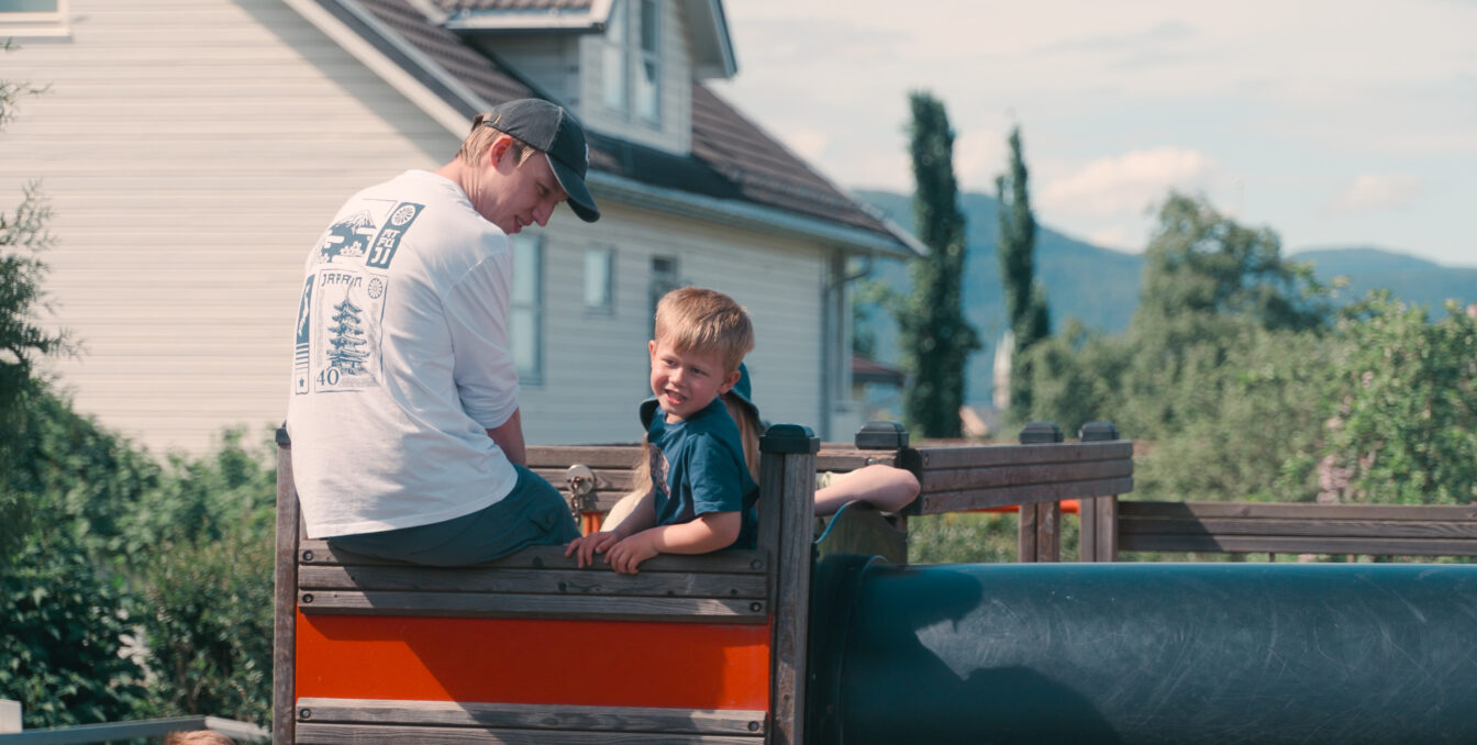 Mannlig barnehageansatt og gutt sitter oppe i et lekestativ på en solfylt dag.