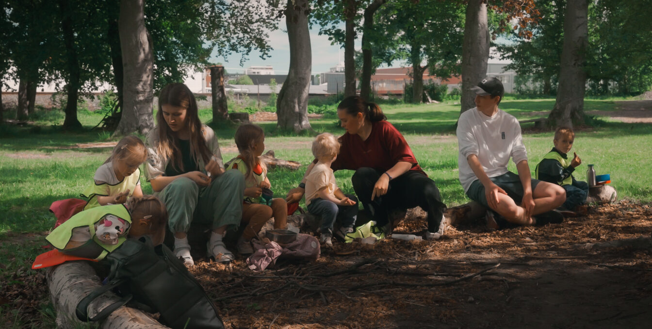 Fire barn og tre voksen sitter på en trestamme i en park og spiser lunsj sammen.