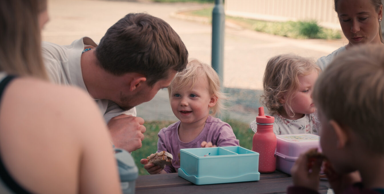Flere barn og voksne sitter å spiser på en benk. En voksen lener seg ned til et av barna. Barnet smiler med en brødskive i hånda.