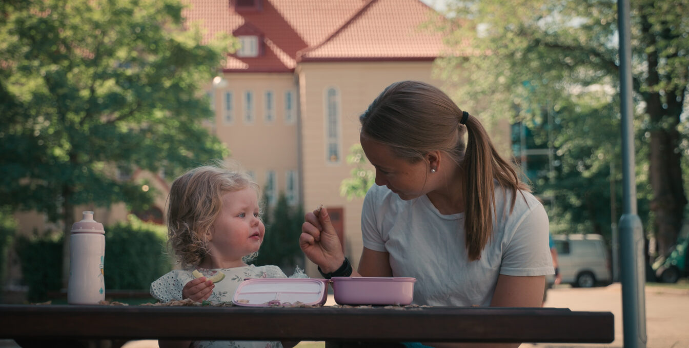 Jente spiser lunsj på en benk sammen med en barnehageansatt. Ser undrende på maten som blir vist til henne.
