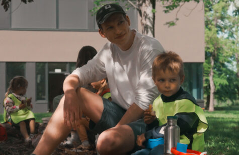 Bilde av en mannlig barnehageansatt og to barn i refleks vester under et tre. De spiser brødskiver og koser seg.