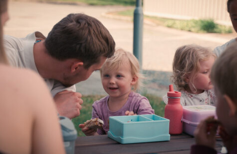 Flere barn og voksne sitter å spiser på en benk. En voksen lener seg ned til et av barna. Barnet smiler med en brødskive i hånda.