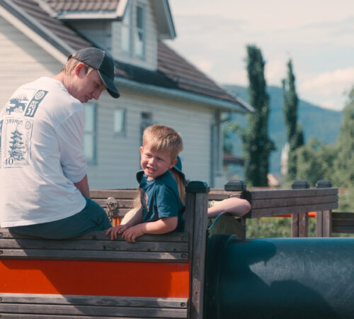 Mannlig barnehageansatt og gutt sitter oppe i et lekestativ på en solfylt dag.