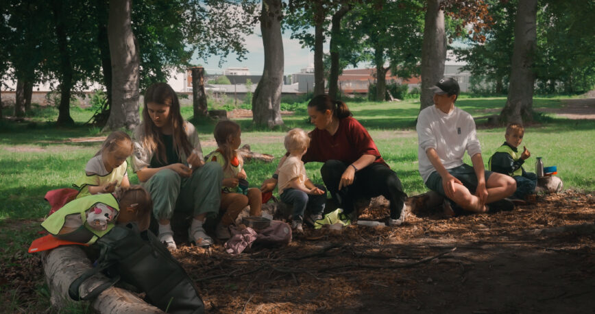 Fire barn og tre voksen sitter på en trestamme i en park og spiser lunsj sammen.