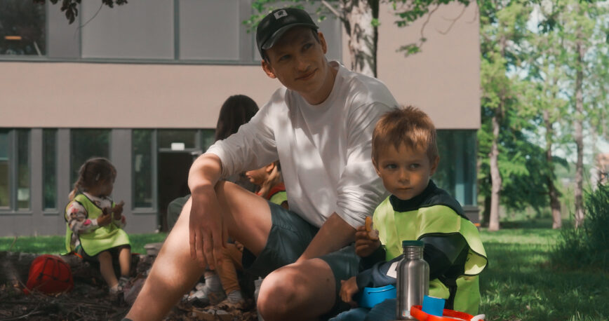 Bilde av en mannlig barnehageansatt og to barn i refleks vester under et tre. De spiser brødskiver og koser seg.