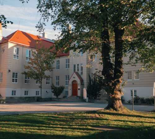 fleretasert lys bygning med rød tak omgitt av trær, inngangsparti med dor, stille campusområde, grønt gress og skygger i ettermiddagssol.