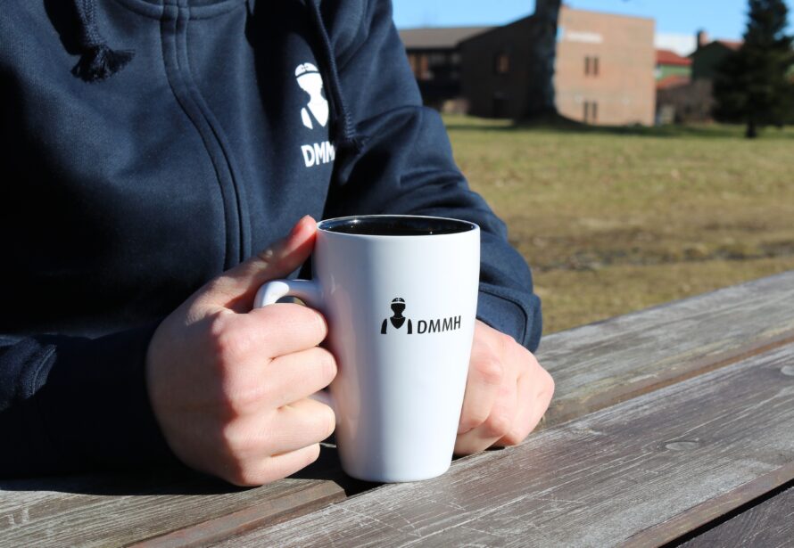 Bilde av en hvit og sort DMMH kopp som blir holdt av en student på et bord.