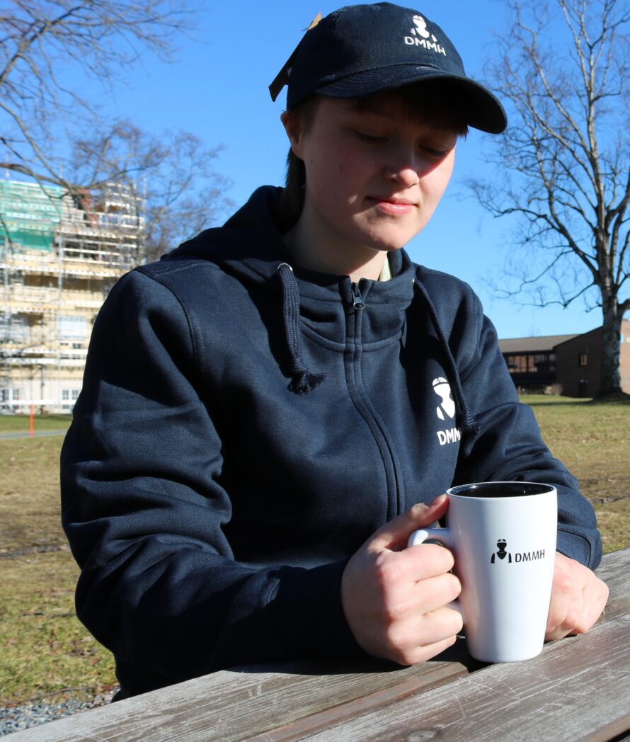 Bilde av en hvit og sort DMMH kopp som blir holdt av en student på et bord.