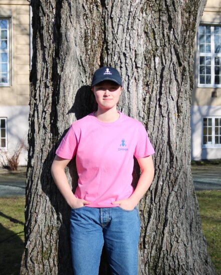 Kvinnelig student med en rosa DMMH t skjorte lener seg mot et tre.