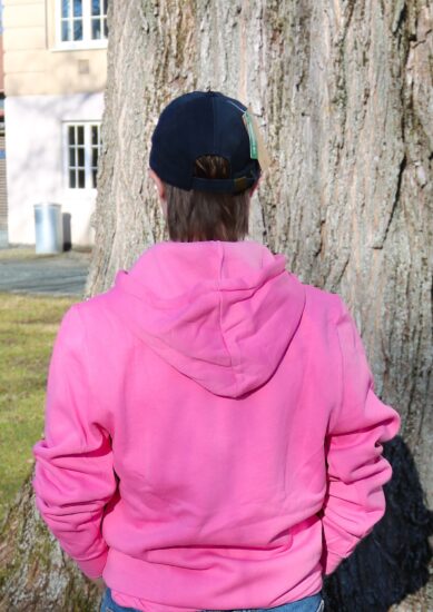Bilde av en student i en rosa DMMH hettegenser foran et tre i parken foran hovedbygget.