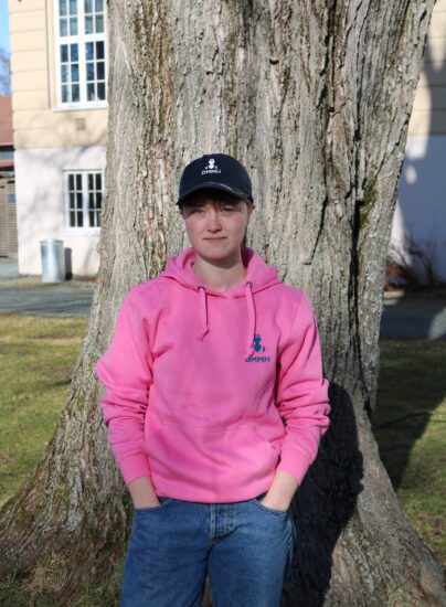 Bilde av en student i en rosa DMMH hettegenser foran et tre i parken foran hovedbygget.