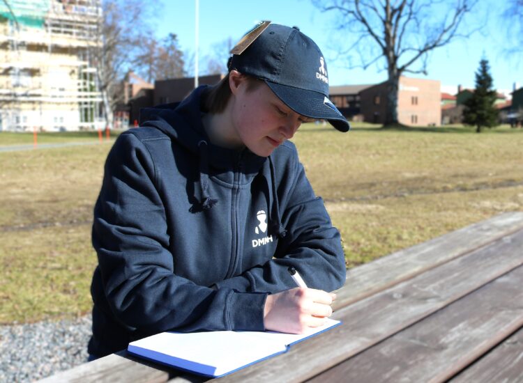 Kvinnelig student skriver i DMMH notatbok foran hovedbygget kledd i DMMH merch.