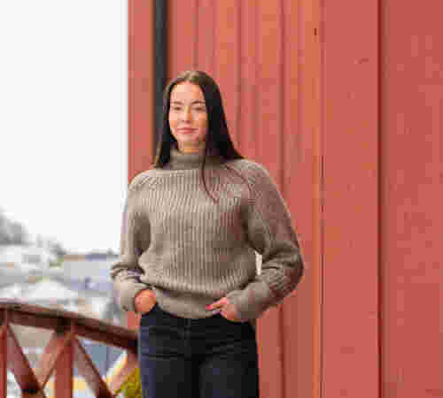 Kvinnelig student i en brun ullgenser står foran en rød låvedør