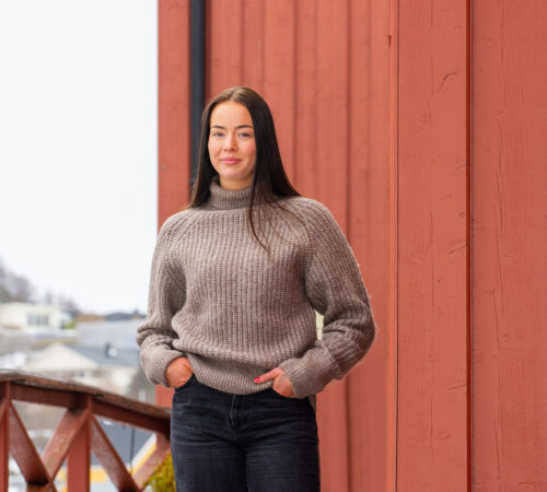 Kvinnelig student i en brun ullgenser står foran en rød låvedør