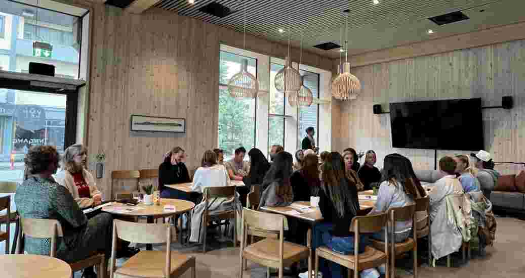 Et stort klasserom der flere studenter sitter rundt lyse bord.