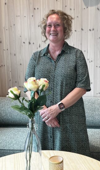 En kvinne står foran et bord med 3 gule roser i en vase.