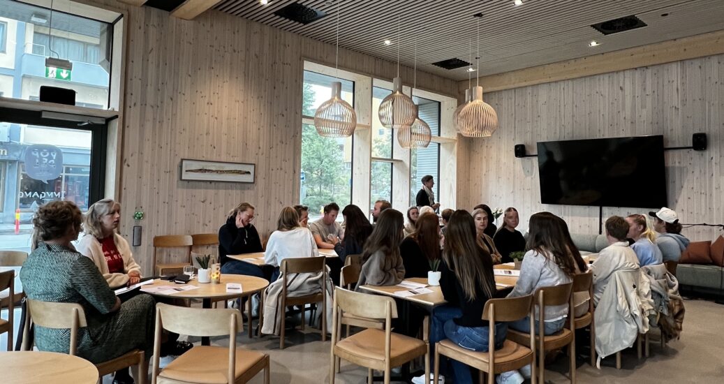 Et stort klasserom der flere studenter sitter rundt lyse bord.