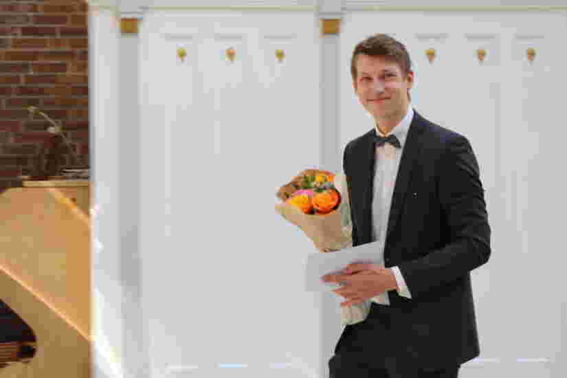 En ung mann i dress med en blomsterbukett i hendene smiler bredt.