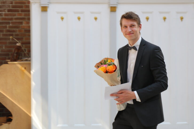 En ung mann i dress med en blomsterbukett i hendene smiler bredt.