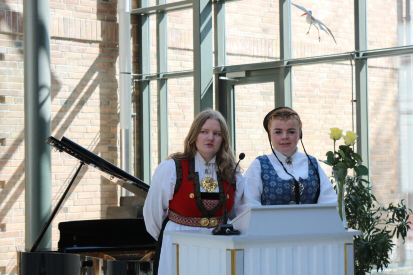 To unge kvinner i bunad står ved en talerstol.