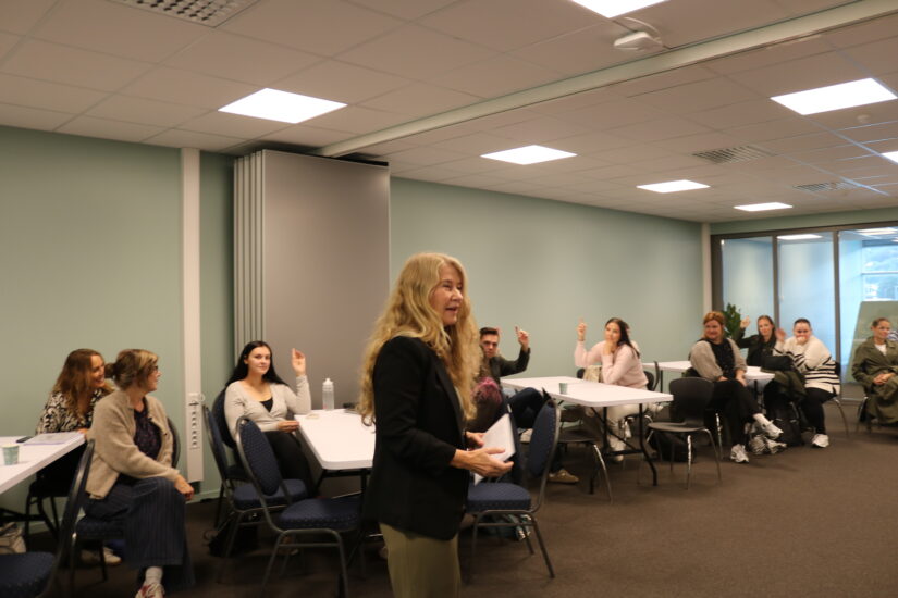 En kvinne står oppreist i et klasserom der mange sitter ved pultene.