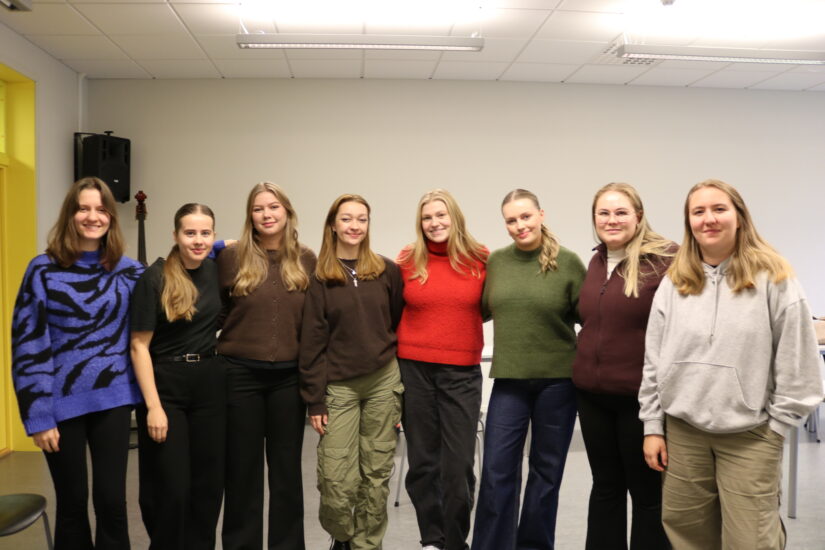 8 unge kvinner står samlet i et rom med en gul dør.