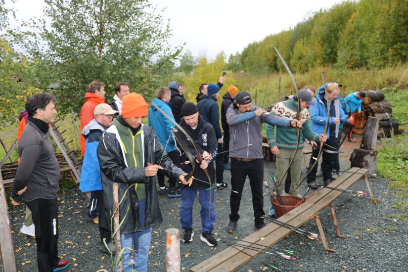 Flere menn står på rad og sikter framover med pil og bue.