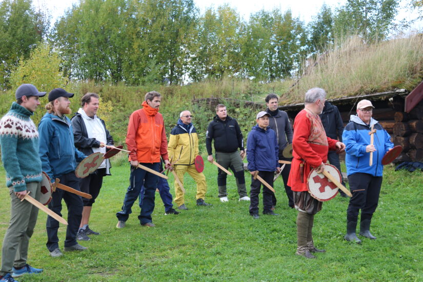 En gruppe menn står på en rekke, de holder tresverd i hendene. En mann i vikingdrakt står med ryggen mot dem.