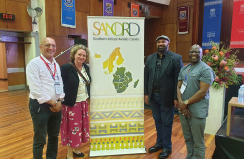 DMMH styrker internasjonale bånd gjennom SANORD-medlemskap. Bilde fra gymsalen på DMMH som viser tre menn og en kvinne foran en plakat for Southern Northern African Senter