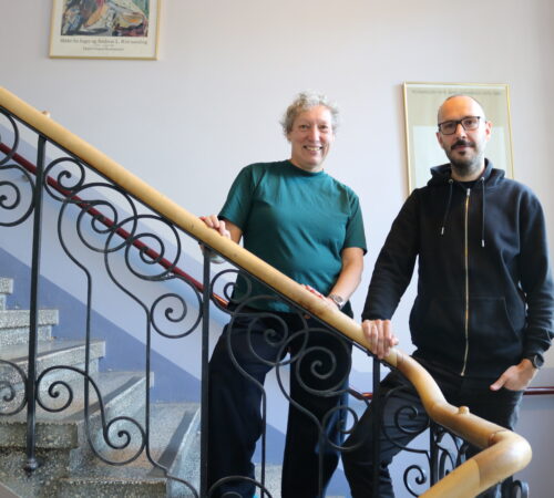 En kvinne og en mann er avbildet stående i en innendørs trappeoppgang med smijernsrekkverk.