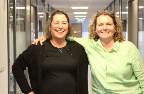 Two woman are photographed standing in a hallway.