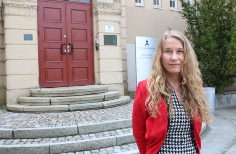 En kvinne med langt krøllete blondt hår står utenfor en bygning med rødt dørlokk i bakgrunnen, iført rød cardigan og sort-hvitt mønsterkjole.