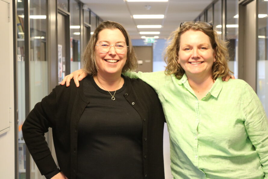 Two woman are photographed standing in a hallway.