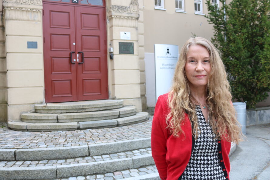 En kvinne med langt krøllete blondt hår står utenfor en bygning med rødt dørlokk i bakgrunnen, iført rød cardigan og sort-hvitt mønsterkjole.