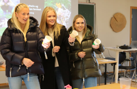 Elever fra videregående fikk prøve studentlivet på DMMH Tre unge personer står sammen i et klasserom og holder papir- og papirkopper, som om de er i en spørrerunde eller lek.
