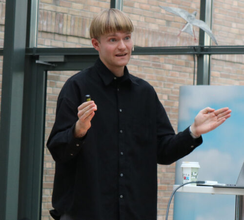 En person med lyst hår som snakker og gestikulerer med hendene, står ved en presentasjonsflate med en kopp og utstyr, bak en glassvegg og murvegg.