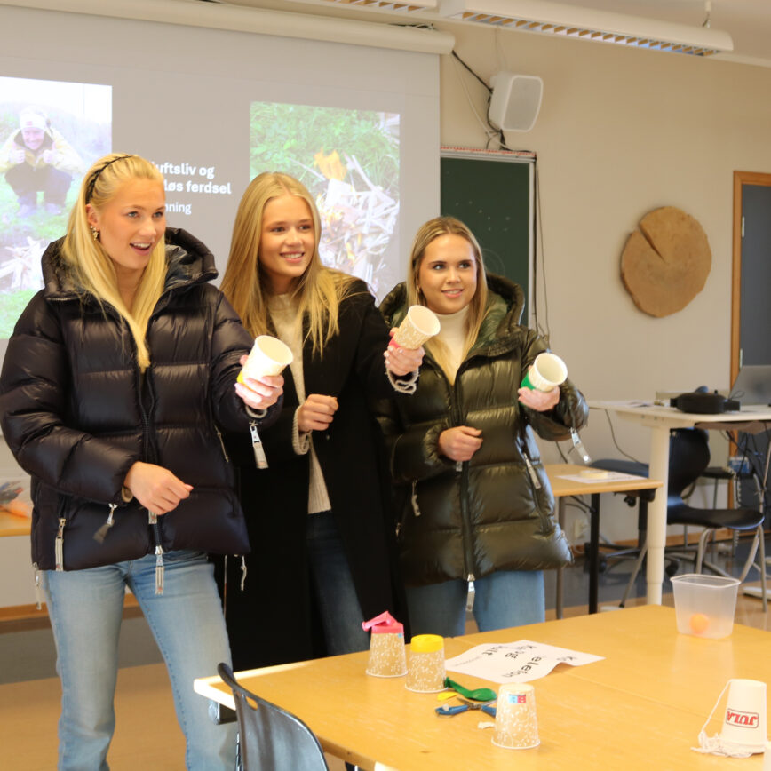 Tre unge personer står sammen i et klasserom og holder papir- og papirkopper, som om de er i en spørrerunde eller lek.