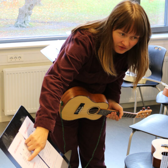 En kvinne med langt brunt hår bøyer seg fremover mens hun holder en liten akustisk gitar og peker på et noteark på et bord i et rom med vindu og stoler.