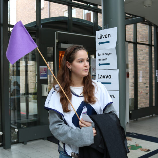 Ung kvinne står inne i bygg med en lilla vimpel i hånden, iført lys-genser og hvit fabric-banner, ved siden av en vegg med informasjonsoppslag.