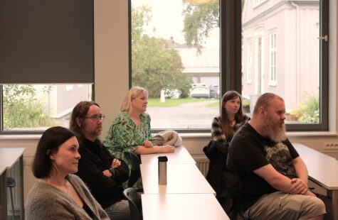 En gruppe fem personer sitter i et lyst klasserom ved en lang rekke bord med vinduer bak dem; utenfor ser man en gate og hus.