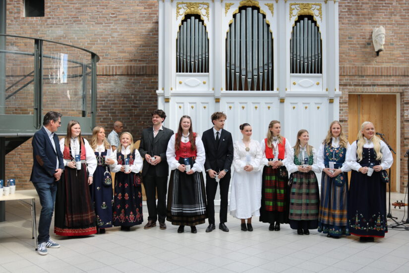 Mange unge mennesker står oppstilt foran et stort orgel, de har fine klær på seg.