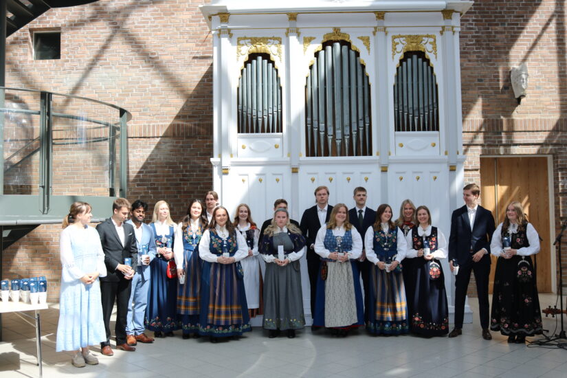 Mange unge mennesker står oppstilt foran et stort orgel, de har fine klær på seg.