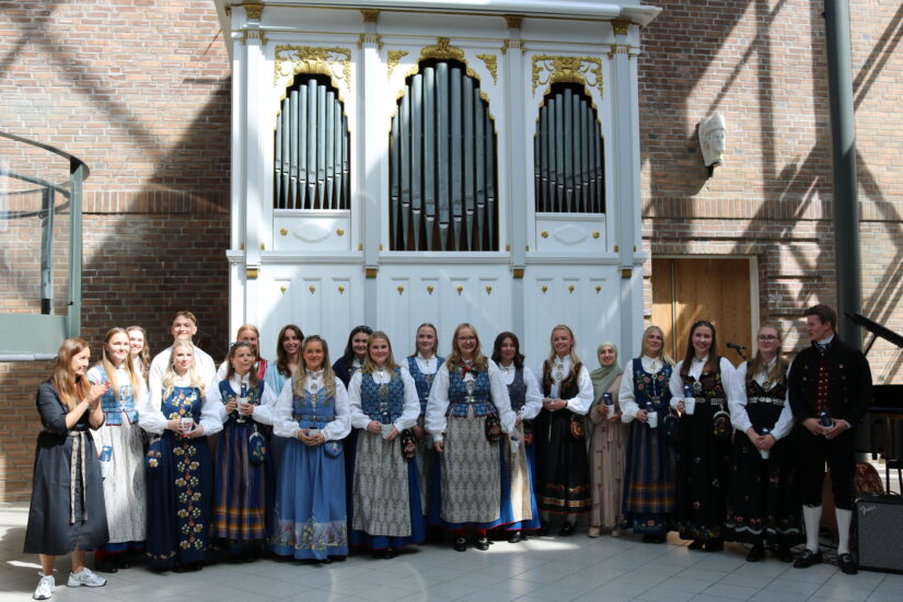 Mange unge mennesker står oppstilt foran et stort orgel, de har fine klær på seg.