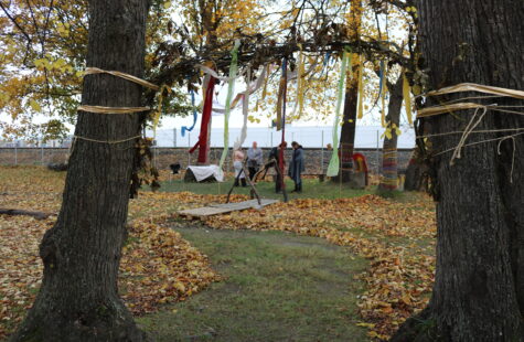 - Artig for både store og små To bålerom med fargede skjerf og papirbånd svirrer mellom trær i en park; flere personer står samlet i bakgrunnen rundt et bord og en lett tegrank, mens løv ligger på bakken.