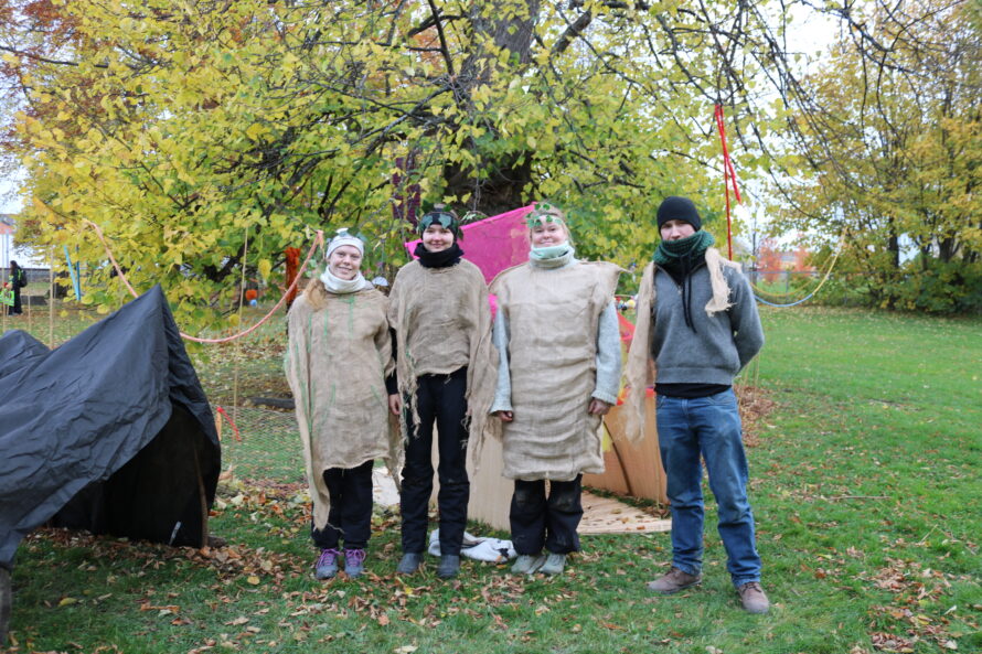 fire personer står sammen i en park omgitt av høstfarger, tre og teltplanter, noen kledd i grove kosedrakter av burlap og skjerf, mens en person har genser og hoderedskap.