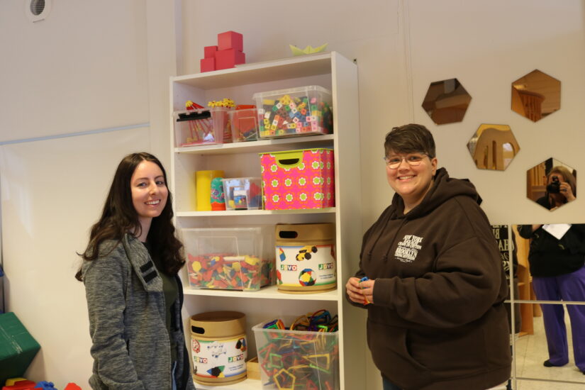 Two young women in front of a bookshelf filled with colourful boxes.