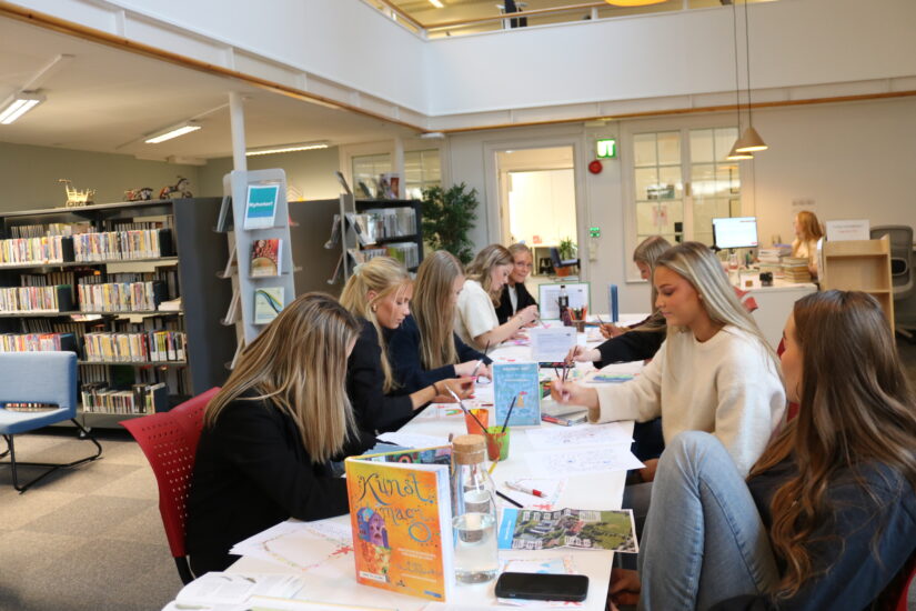 En gruppe ungdommer sitter rundt et langbord. Det er barnebøker og malingssutstyr på bordet.