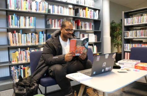 En mann leser en bok med en laptop foran seg. Bak er det mange bokhyller.