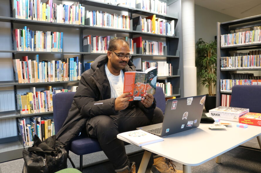 En mann leser en bok med en laptop foran seg. Bak er det mange bokhyller.